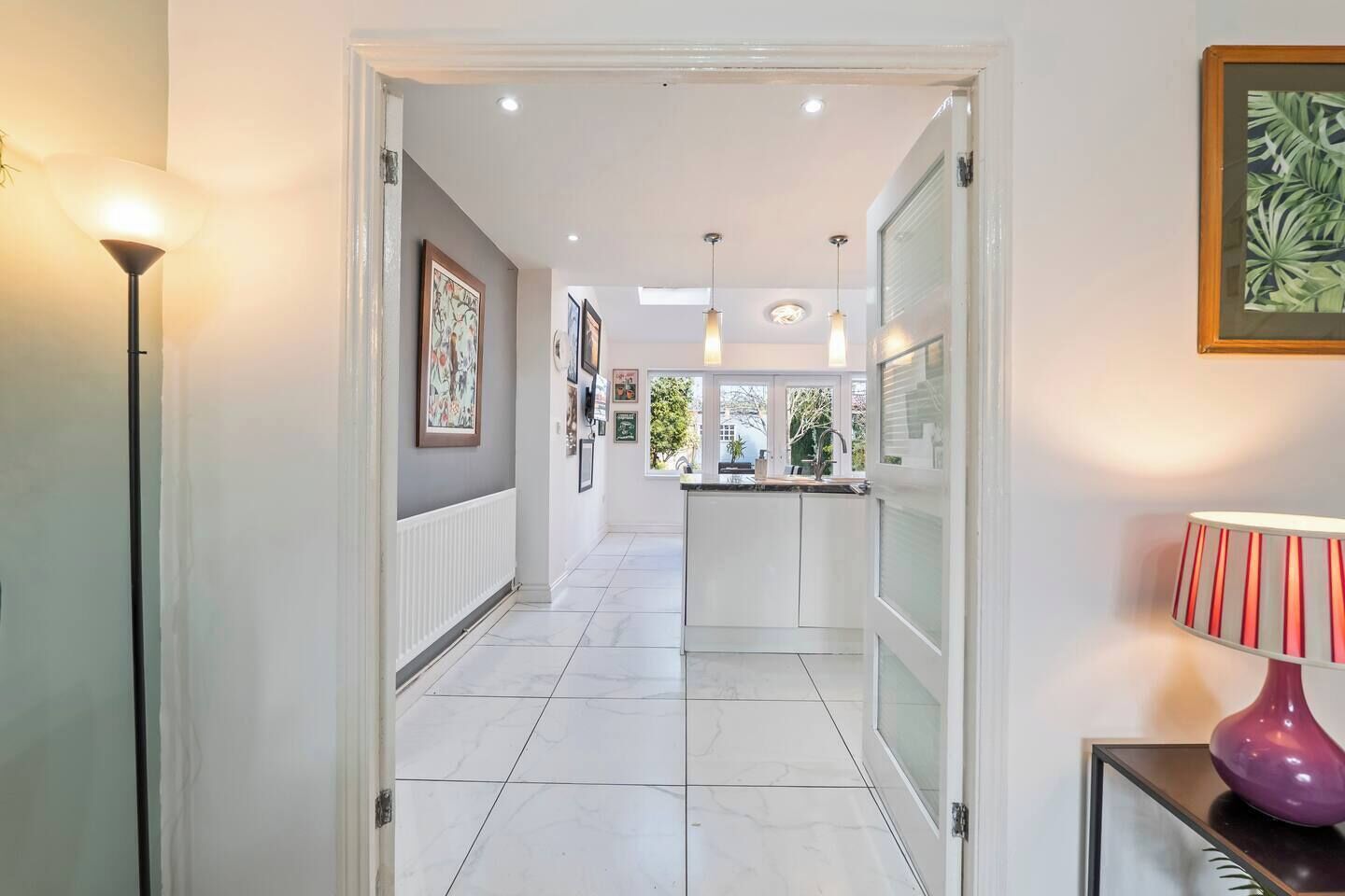 A hallway with a kitchen area, white tiled flooring, and pendant lighting.