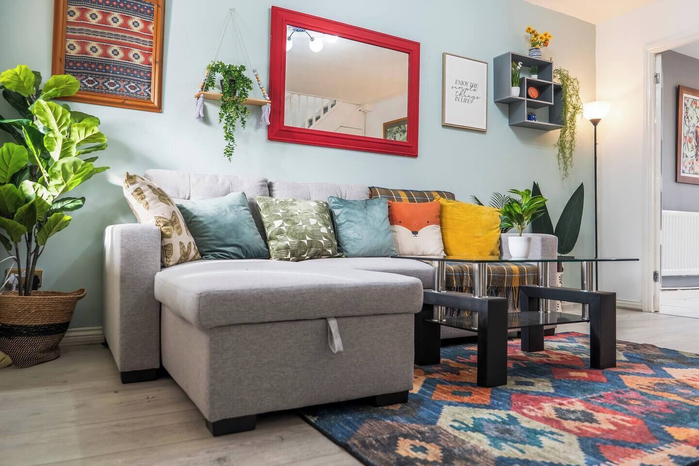 Een woonkamer met een grijze bank, kleurrijke kussens, een spiegel met rode lijst en een vloerkleed met een patroon.