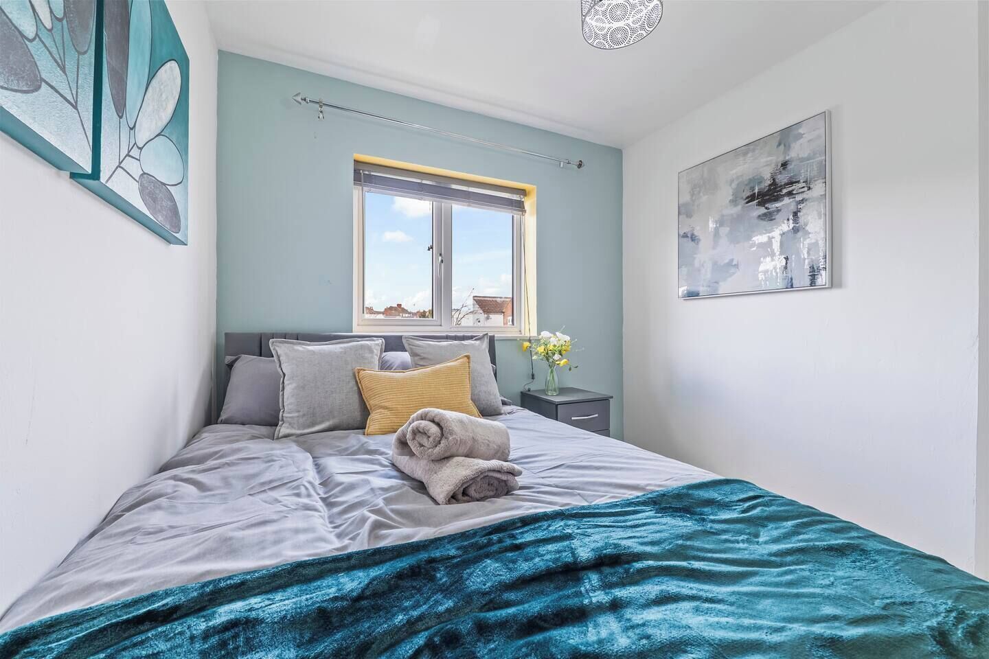A bedroom with a bed, pillows, a window, a nightstand, a vase with flowers, and a painting on the wall.