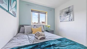 A bedroom with a bed, pillows, a window, a nightstand, a vase with flowers, and a painting on the wall.