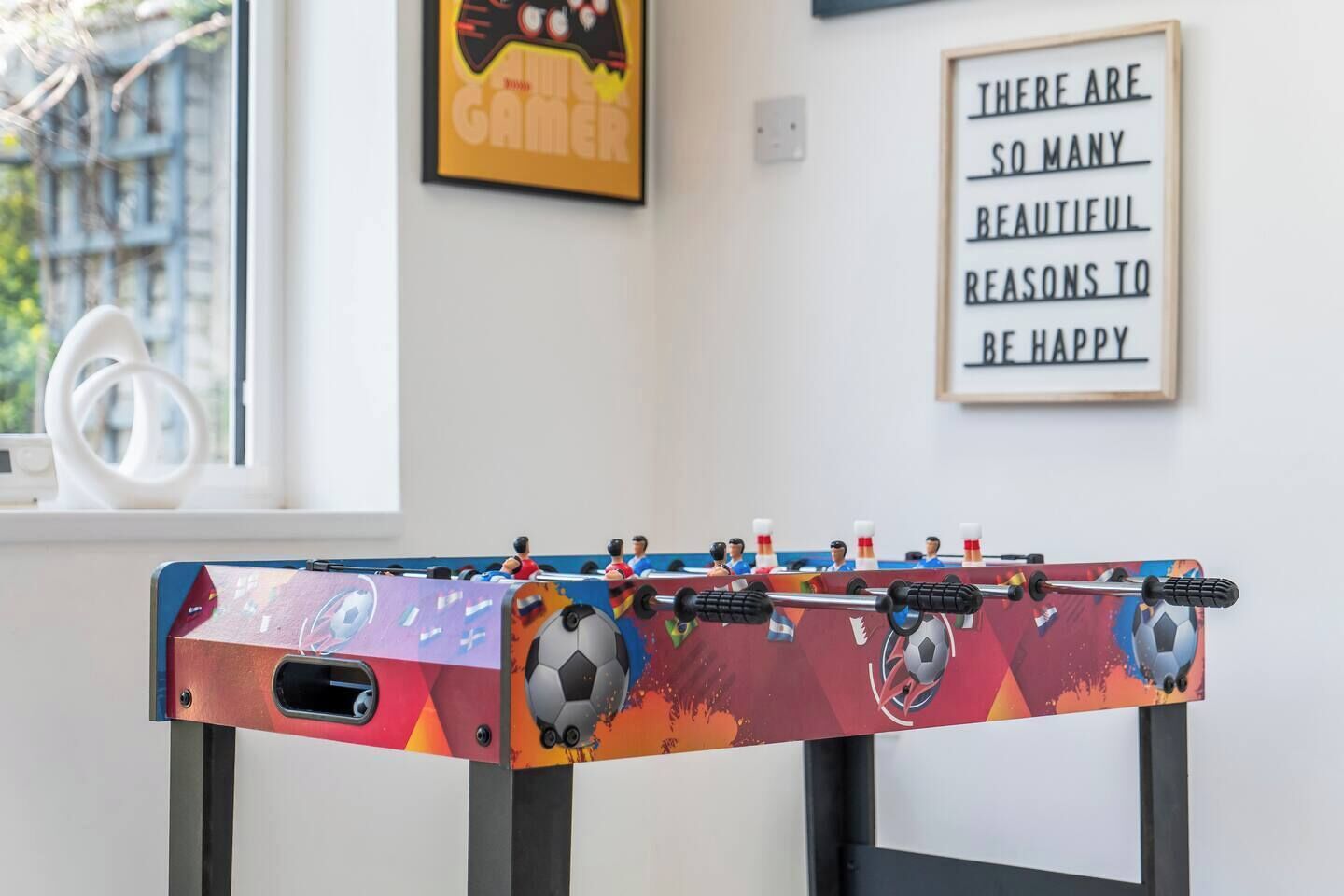 Een tafelvoetbaltafel met een kleurrijk ontwerp, een inspirerende quote op de muur en een raam met uitzicht op een gebouw.