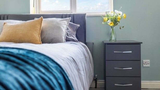 Quarto com cama, almofadas, mesa de cabeceira com um vaso de flores e janela com vista para o céu.