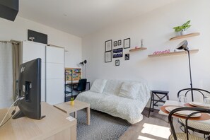 Ein modernes Wohnzimmer mit Sofa, Fernseher, Schreibtisch und Stuhl. An den Wänden hängen Bilder in Rahmen und auf einem Regal steht eine kleine Pflanze.
