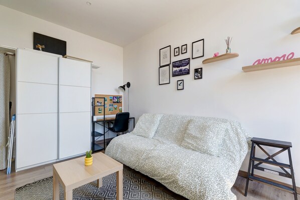 Ein Zimmer mit einer gepolsterten Couch mit Muster, einem kleinen Tisch mit Pflanze, einem Schreibtisch mit Stuhl und einer Pinnwand mit Fotos.