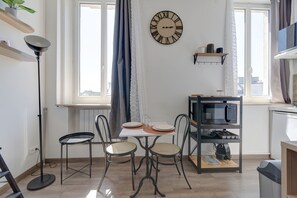 A dining area with a table set for two, a clock on the wall, and a kitchen area with a microwave. - L'Escale Rémoise - À 600 m de la Porte de Mars (Reims)