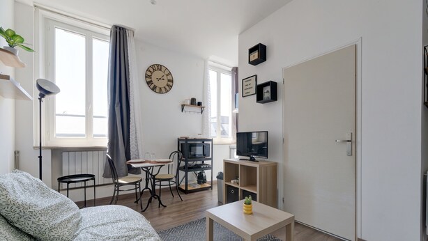 A modern living room with a sofa, a dining table with chairs, a TV, and a clock on the wall. - L'Escale Rémoise - À 600 m de la Porte de Mars (Reims)
