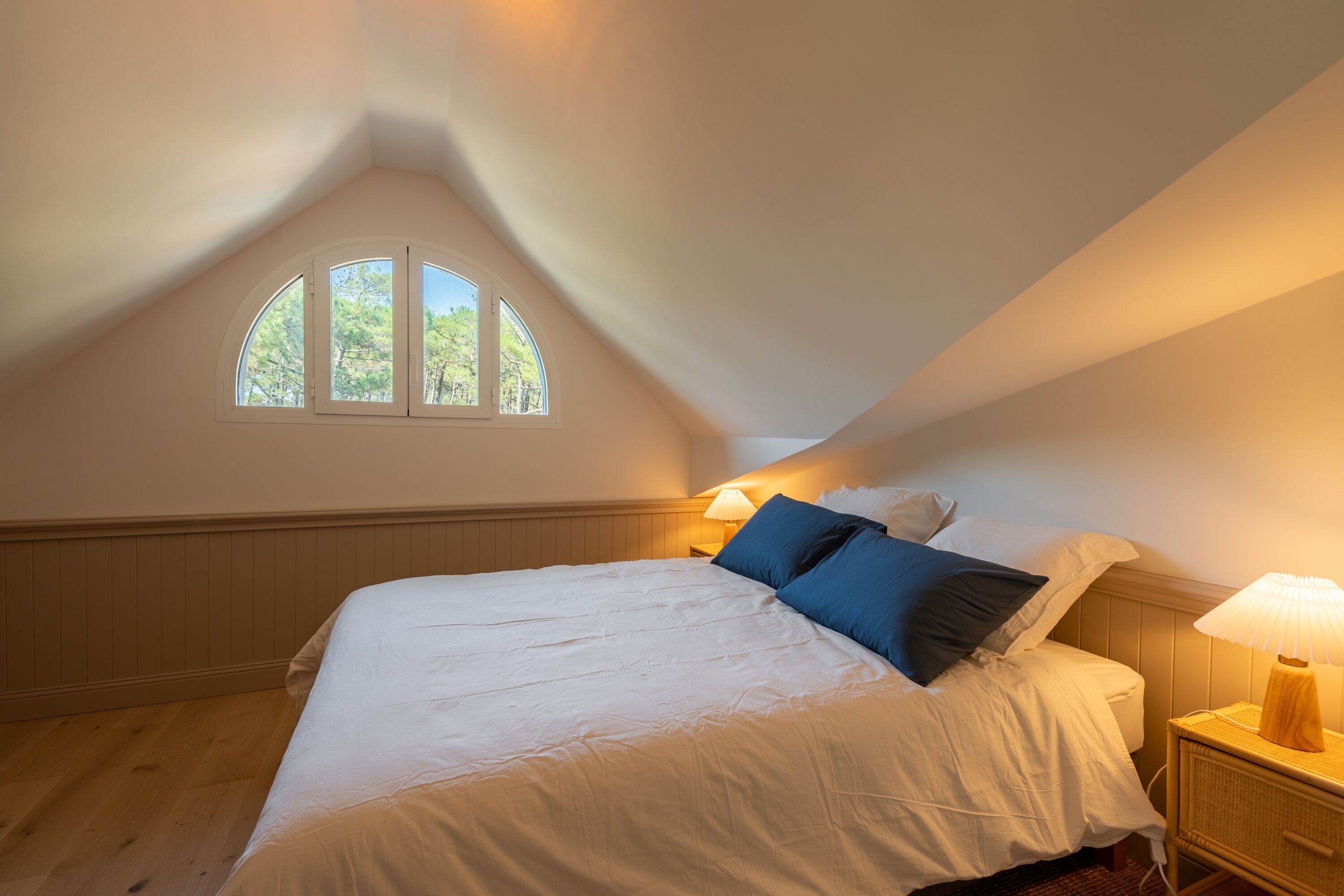 A bedroom with a sloped ceiling, a large bed, two bedside lamps, and a window with a view of trees.