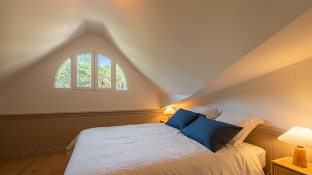 Una camera da letto con soffitto spiovente, un letto grande, due abat-jour e una finestra con vista sugli alberi.