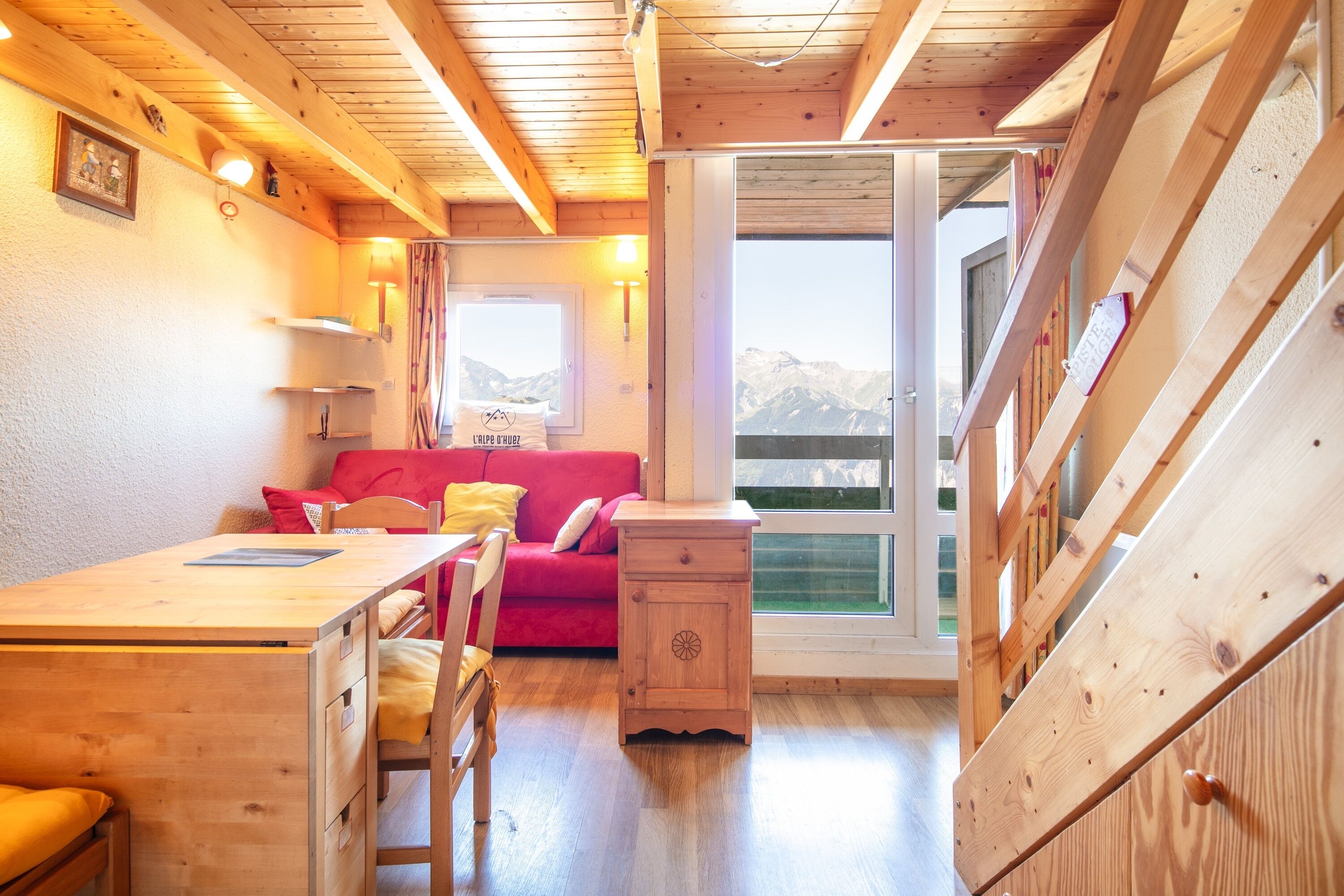 Ein gemütliches Blockhaussinnere mit einem Holzespeisetisch, einem roten Sofa und Bergblick durch ein großes Fenster.