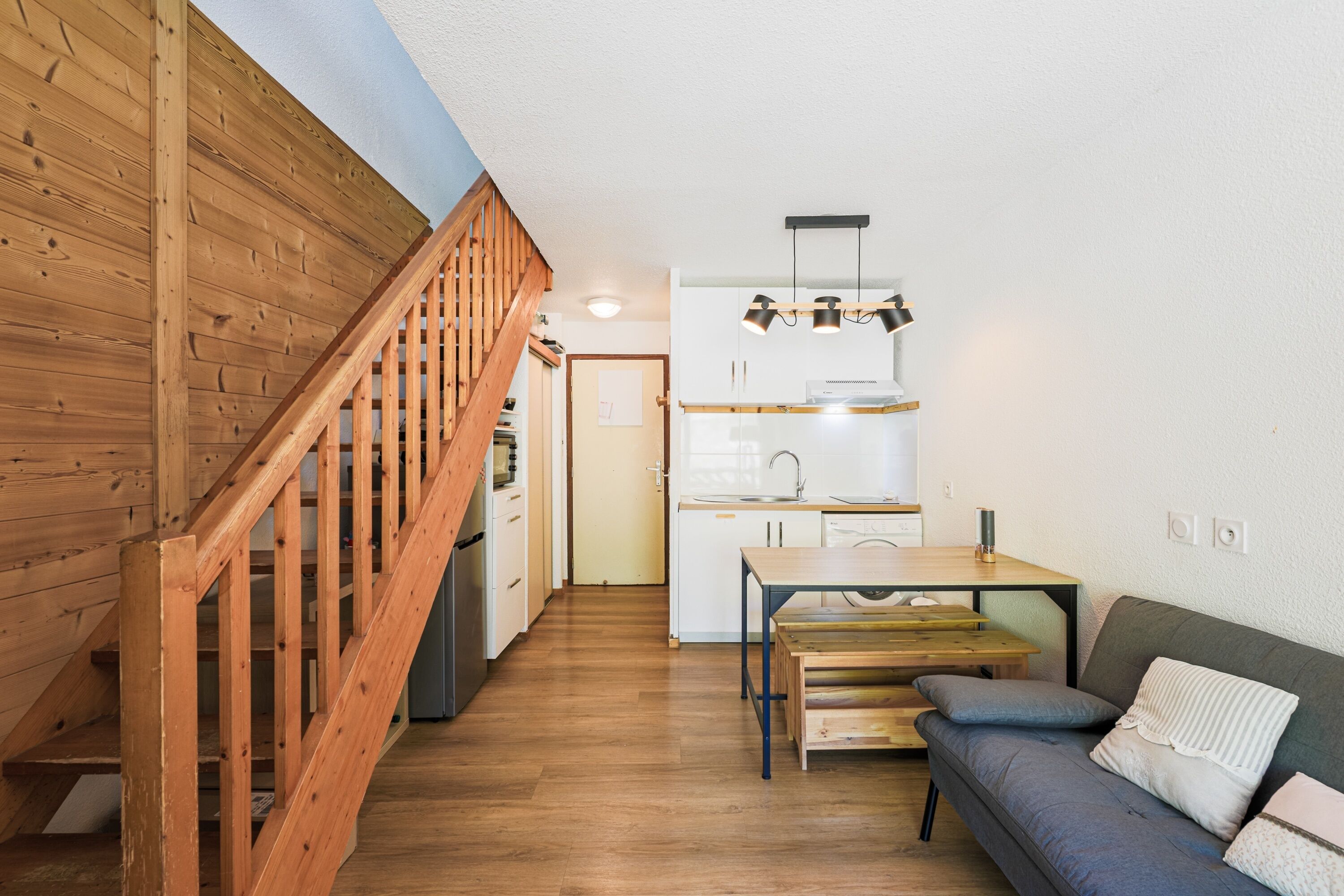 Une cuisine moderne avec un escalier en bois, un coin repas avec une table et des bancs, et un canapé gris agrémenté de coussins.