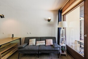 A cozy room with a grey sofa, a wooden side table, a lamp, and a view through a large window. - Chez Joséphine - Station de ski accessible à pied (Gresse-en-Vercors)