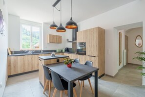 A modern kitchen with wooden cabinets, a dining table with chairs, and pendant lighting. - Gîte La Petite Récré - Au pied des Hautes Vosges (Plainfaing)