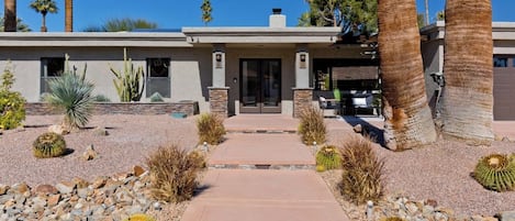 Una casa moderna con entrada pavimentada, rodeada de plantas de desierto y palmeras.