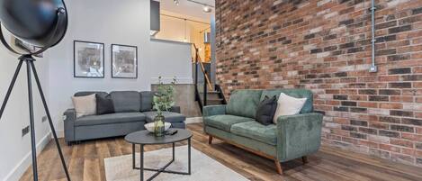 A modern living room with a brick wall, a grey sofa, a green sofa, a round coffee table, and a floor lamp.