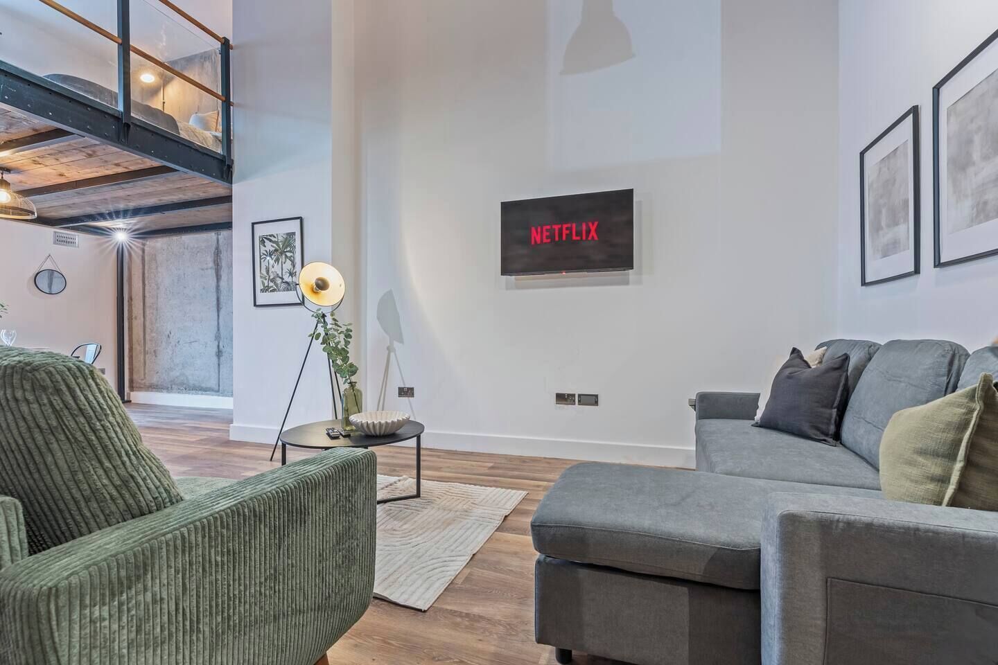 Uma sala de estar moderna com um sofá cinza, uma poltrona verde, um abajur de chão e uma TV na parede exibindo 'NETFLIX'.