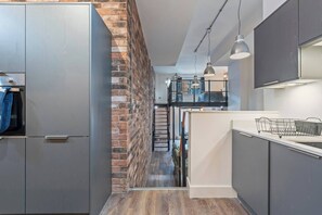Een moderne keuken met grijze kastjes, een accentmuur van baksteen, en een entresol met een trap.