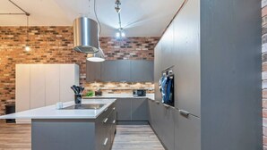 Uma cozinha moderna com uma ilha central, armários cinzentos, parede de tijolos e iluminação pendente.