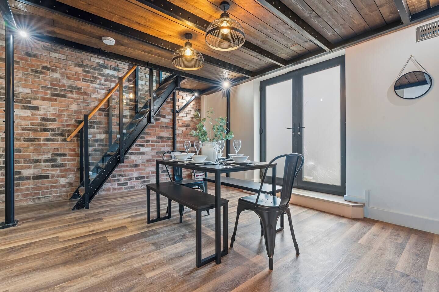 Comedor con pared de ladrillo, suelo de madera, una escalera e iluminación colgante.