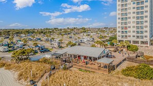 Zona costera con aparcamiento, vehículos recreativos, un restaurante frente al mar y un edificio alto al fondo.