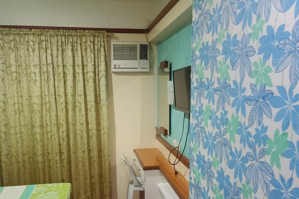 A room with a bed, a desk, a chair, an air conditioning unit, and a wall-mounted television.