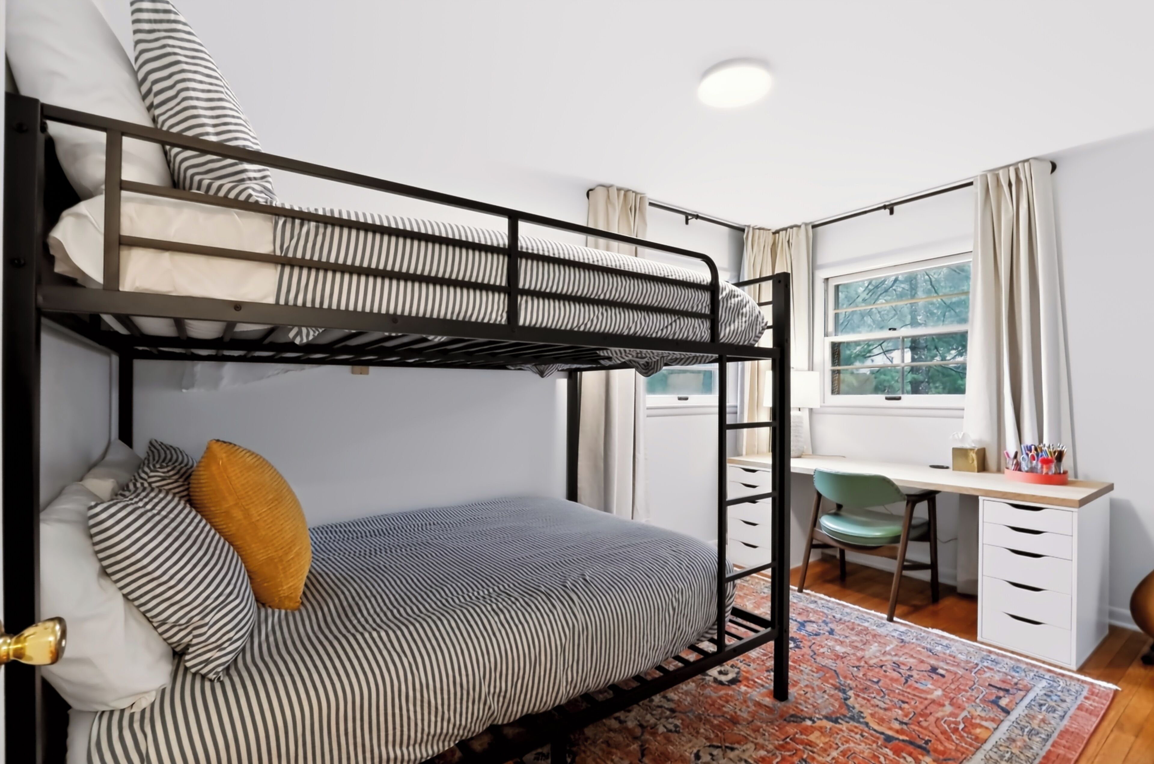 A bunk bed with striped bedding, a desk with a chair, a window with curtains, and a patterned rug.