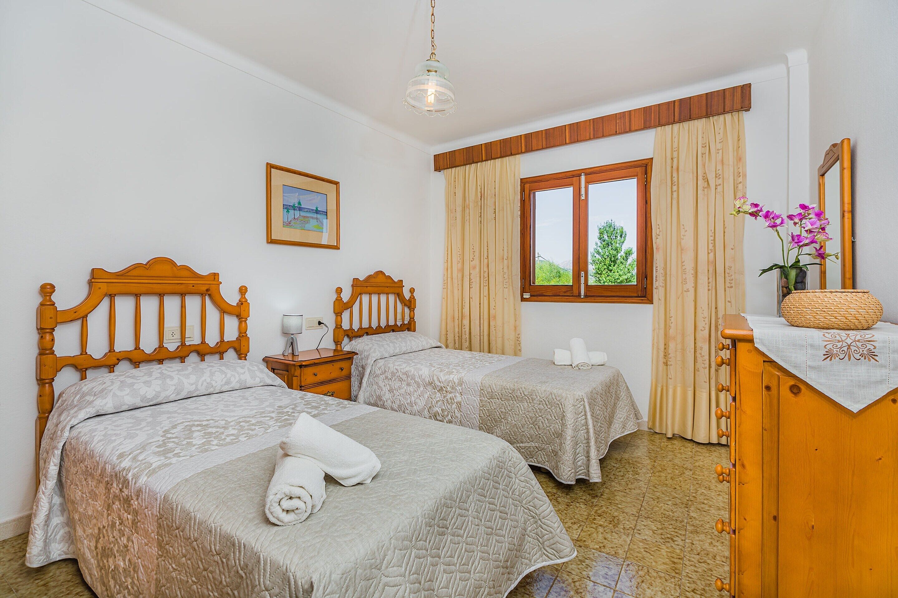 Habitació d'hotel amb dos llits, un capçal de llit de fusta, una taula de nit amb una làmpada, una finestra amb cortines, un quadre encadrejat a la paret i un moble d'emmagatzematge de fusta amb un ram de flors.