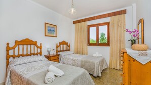 A hotel room with two beds, a wooden headboard, a bedside table with a lamp, a window with curtains, a framed picture on the wall, and a wooden dresser with a vase of flowers. - Elegant Pool Villa in Alcudia (Alcúdia)