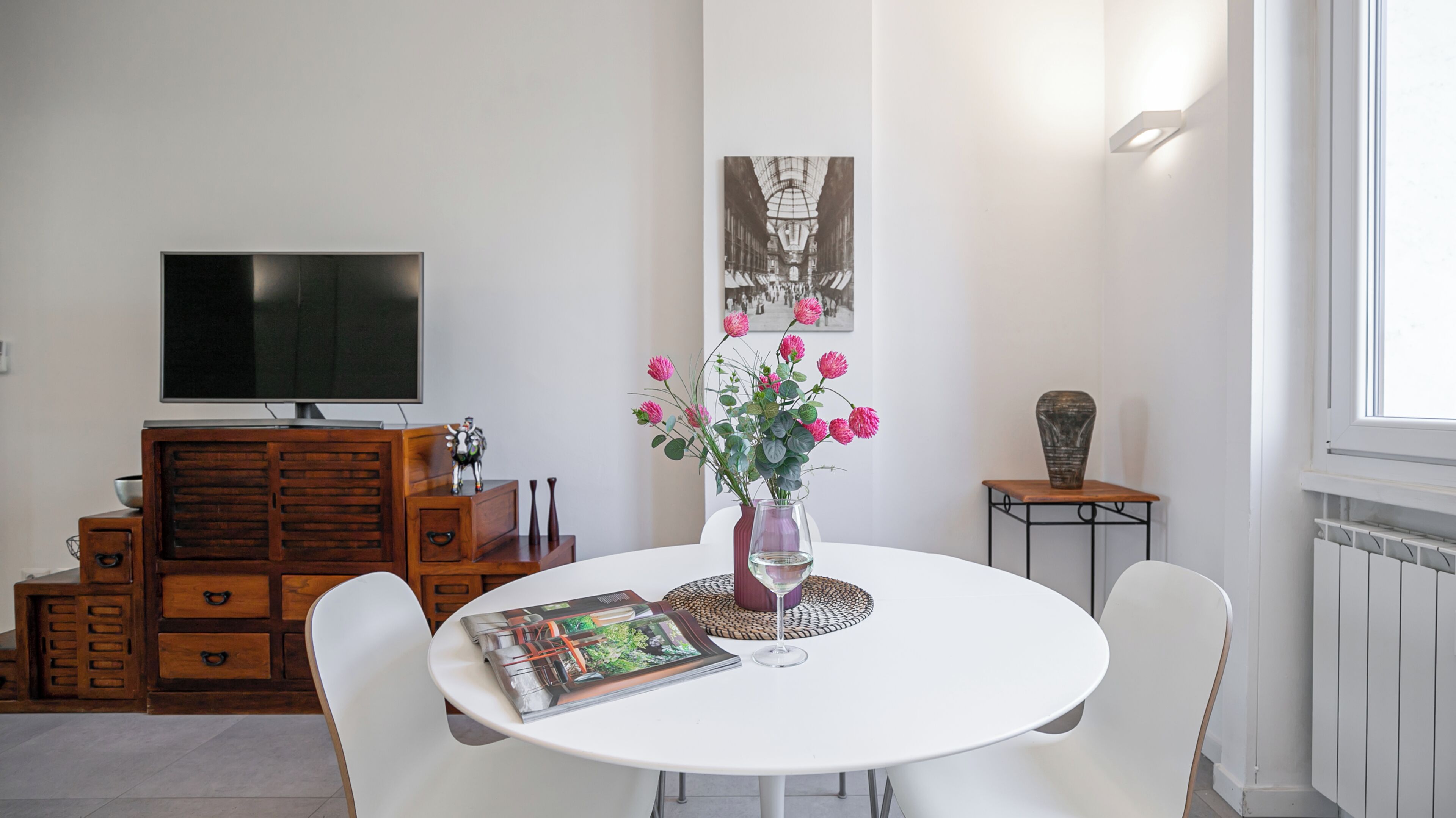A dining area with a round table, white chairs, a vase with pink flowers, and a television on a wooden cabinet.
