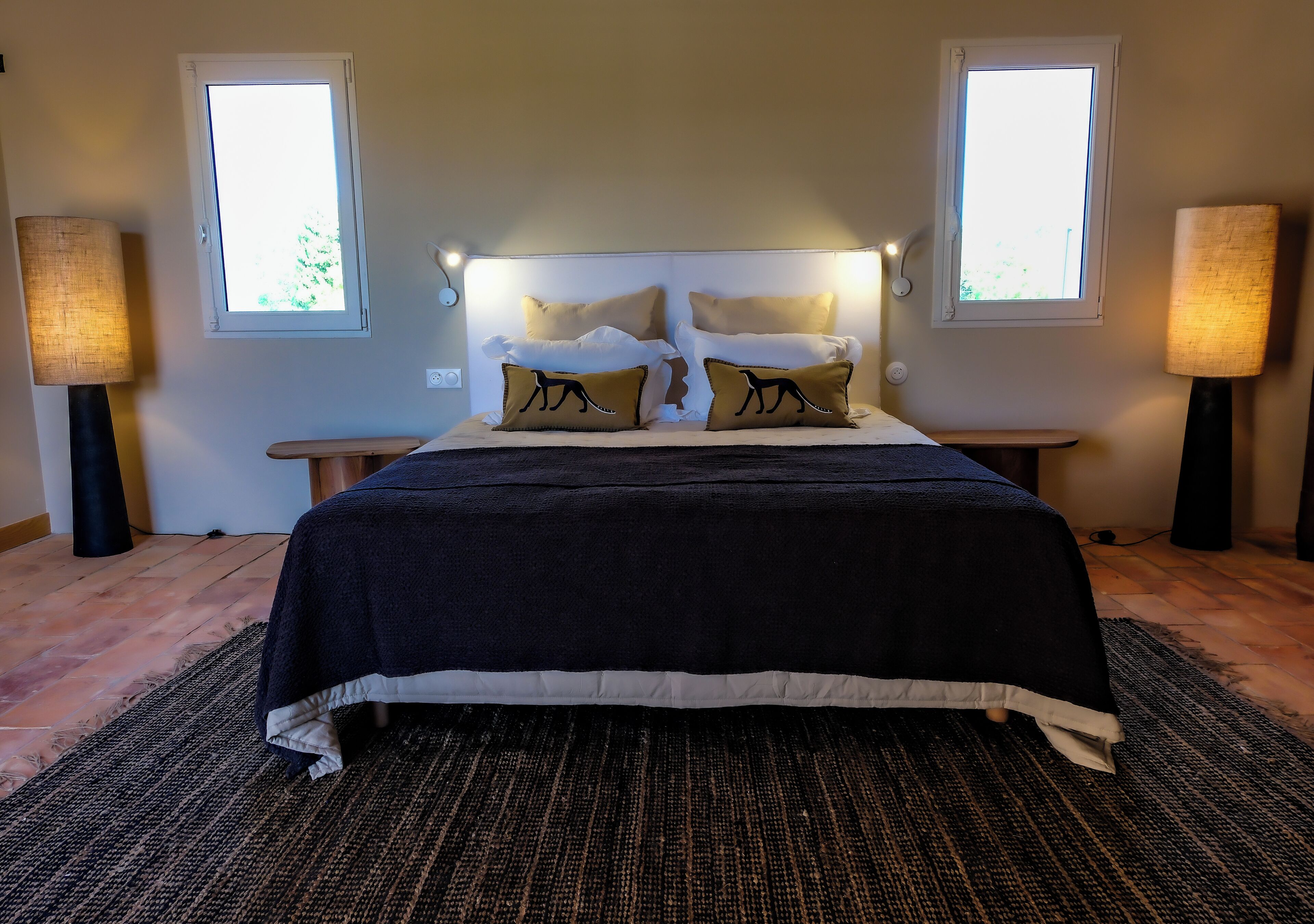 Una cama con un edredón oscuro, dos mesitas de noche con lámparas y dos ventanas con luz natural.