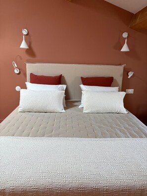 Una cama con cabecera color beige, ropa de cama blanca y dos almohadas rojas.