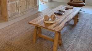 A rustic interior with exposed wooden beams, a wooden table with decorative items, a painting of a desert landscape, and a chair with a cushion labeled 'AMOUR'. - Farmhouse in Provence (Pernes-les-Fontaines)
