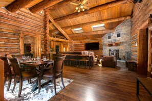 A log cabin interior with a dining area, stone fireplace, and wooden ceiling beams.