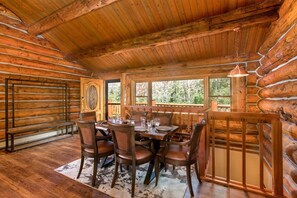A log cabin interior with a dining table set for four, wooden walls, and a large window offering a view of trees.