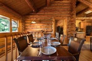 A wooden log cabin interior with a dining table set for four, chairs, a kitchen area, and a large window.