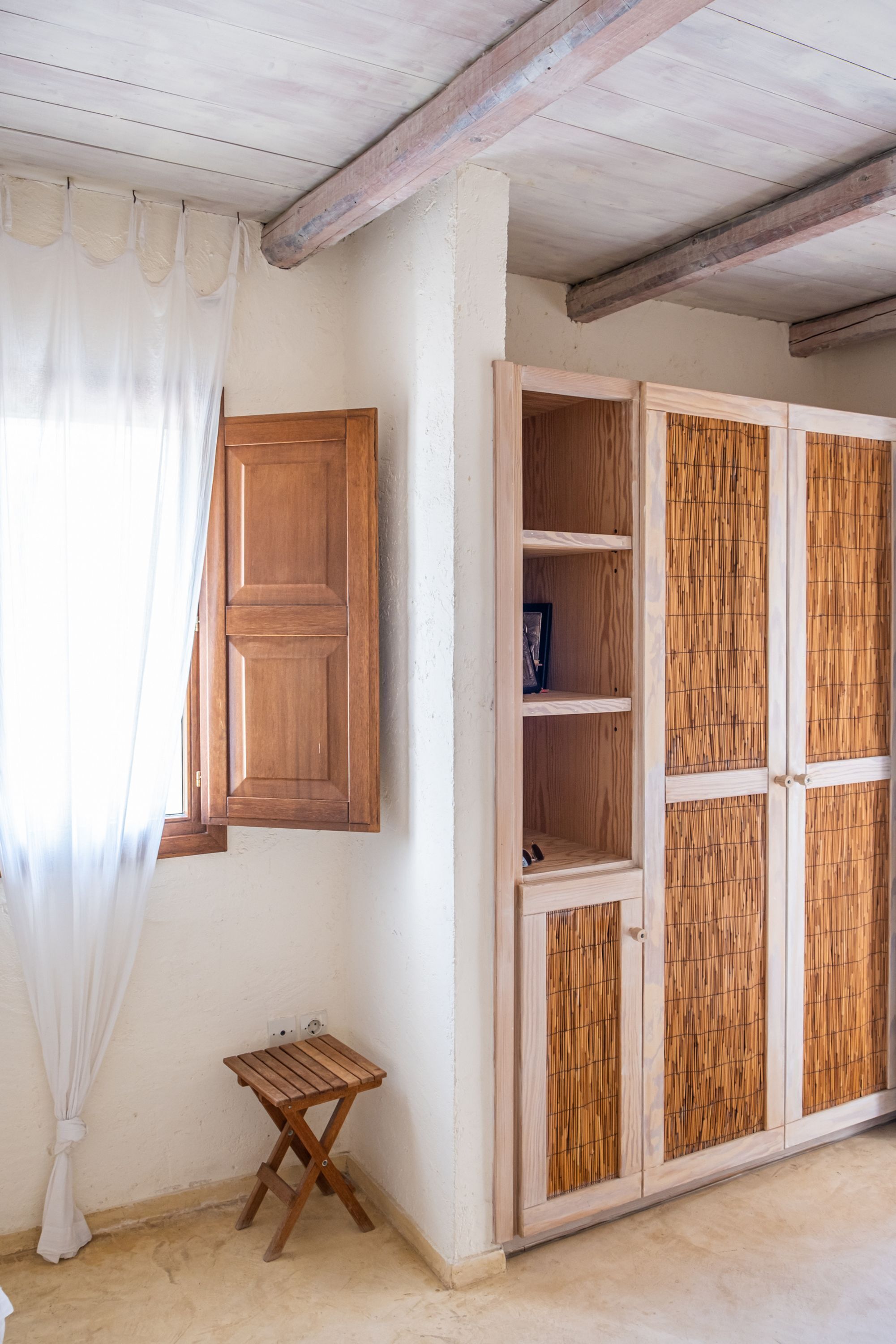 Ein Zimmer mit einem Holfischschrank, einem kleinen Holzschemel und einem Fenster mit weißem Vorhang.