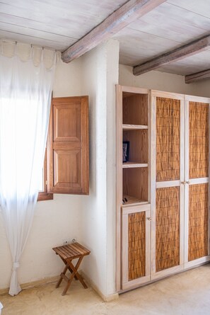 A room with a wooden wardrobe, a small wooden stool, and a window with a white curtain. - Zephyros | Pool & Tennis (Koundouros)