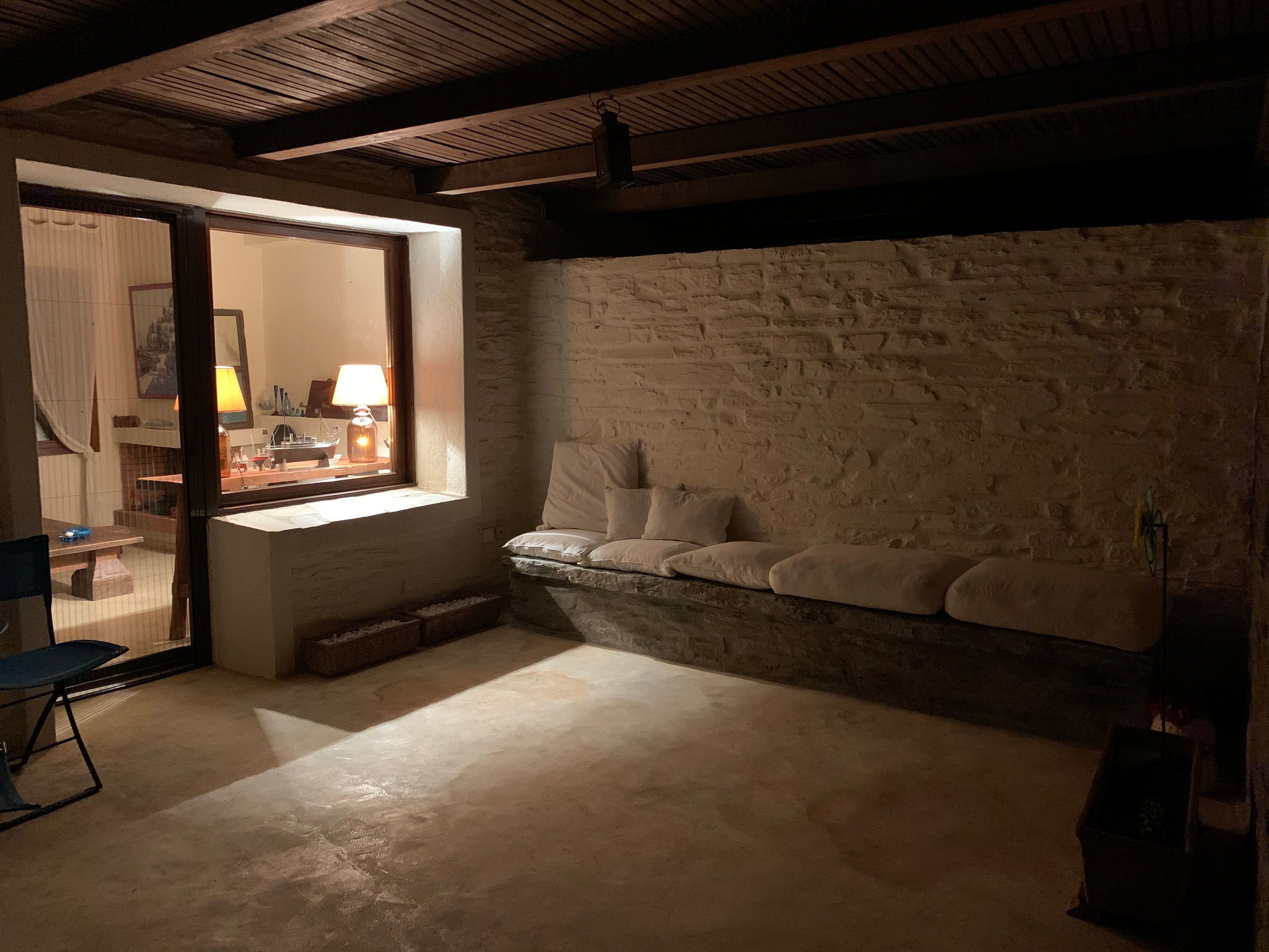 A room with a stone wall, a cushioned bench, a table with a lamp, and a view through an open door.