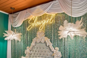 Une chambre avec une tête de lit capitonnée, des décorations en plumes blanches et un néon qui affiche le mot « Showgirl ».