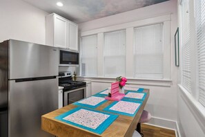 Eine kleine Küche mit einem Edelstahl-Kühlschrank, einer Mikrowelle und einem Herd. Ein Holztisch mit einer blau-rosa Tischdecke ist ebenfalls vorhanden.