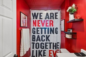 Ein Badezimmer mit roter Wand, einer weißen Tür und einem Handtuch, das an der Tür hängt. Ein Schild mit dem Text 'WE ARE NEVER GETTING BACK TOGETHER LIKE EVER' ist an der Wand angebracht.