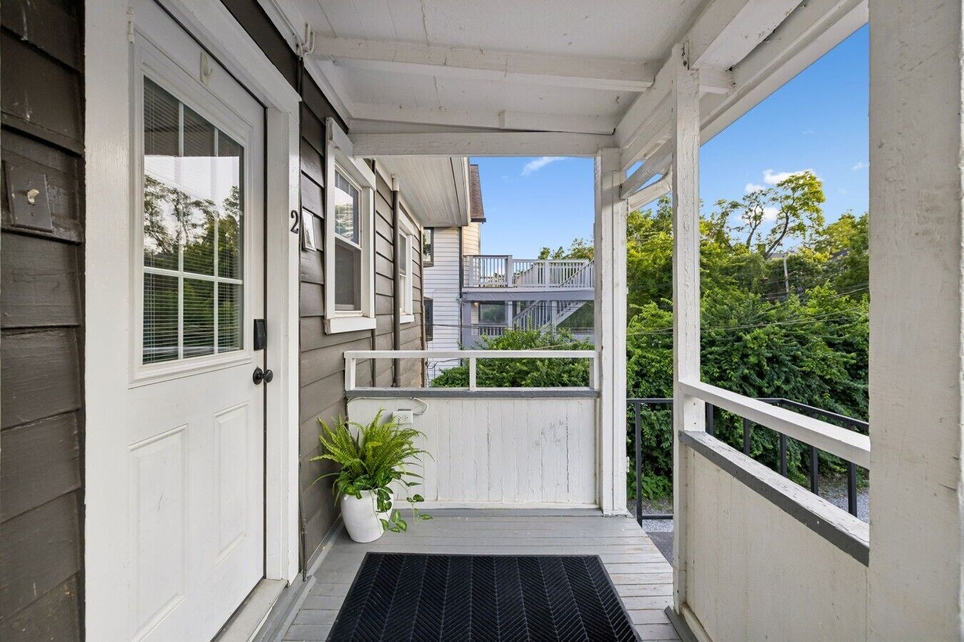 Eine überdachte Veranda mit einer schwarzen Matte, einem Blumentopf und Blick auf ein Nachbargebäude.
