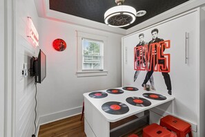Ein Zimmer mit weißer Wand, einem Fenster, einer Deckenleuchte, einem an der Wand befestigten Fernseher und einem Tisch mit Schallplatten.