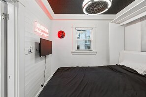 Une chambre à coucher avec un lit, un téléviseur fixé au mur, une fenêtre et un panneau lumineux au néon.