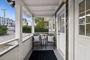 A covered porch with two chairs and a small table, a door to the right, and a view of the street and buildings in the background. - Backwoods Barbie Suite | Sleeps 10 on Music Row (Nashville)