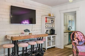 A bar area with stools, a television mounted on a white brick wall, and a kitchen in the background. - Backwoods Barbie Suite | Sleeps 10 on Music Row (Nashville)