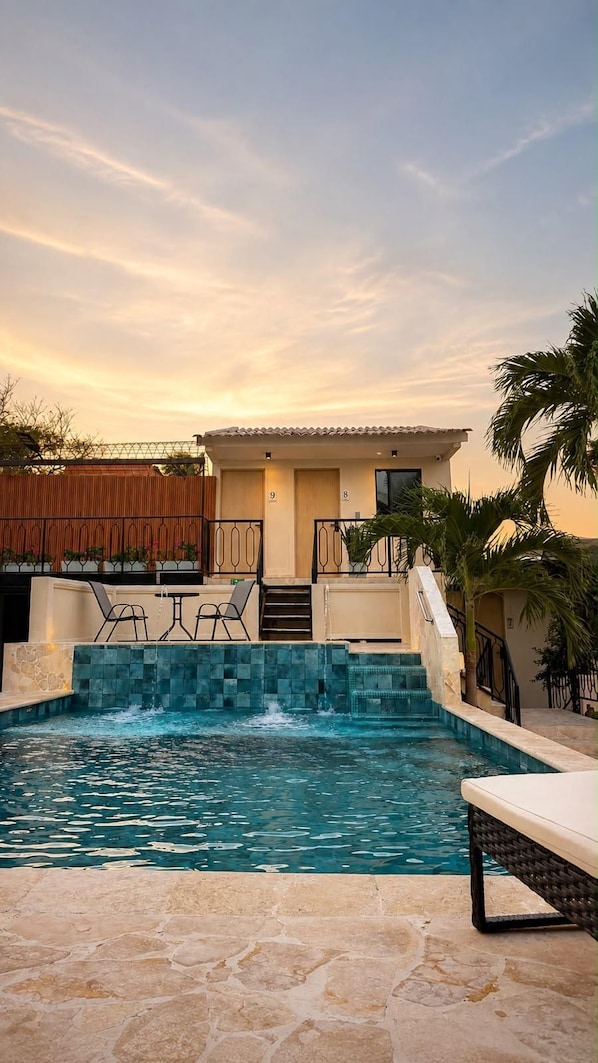 A swimming pool with a stone deck, a lounge area with chairs, and a building with a tiled roof.