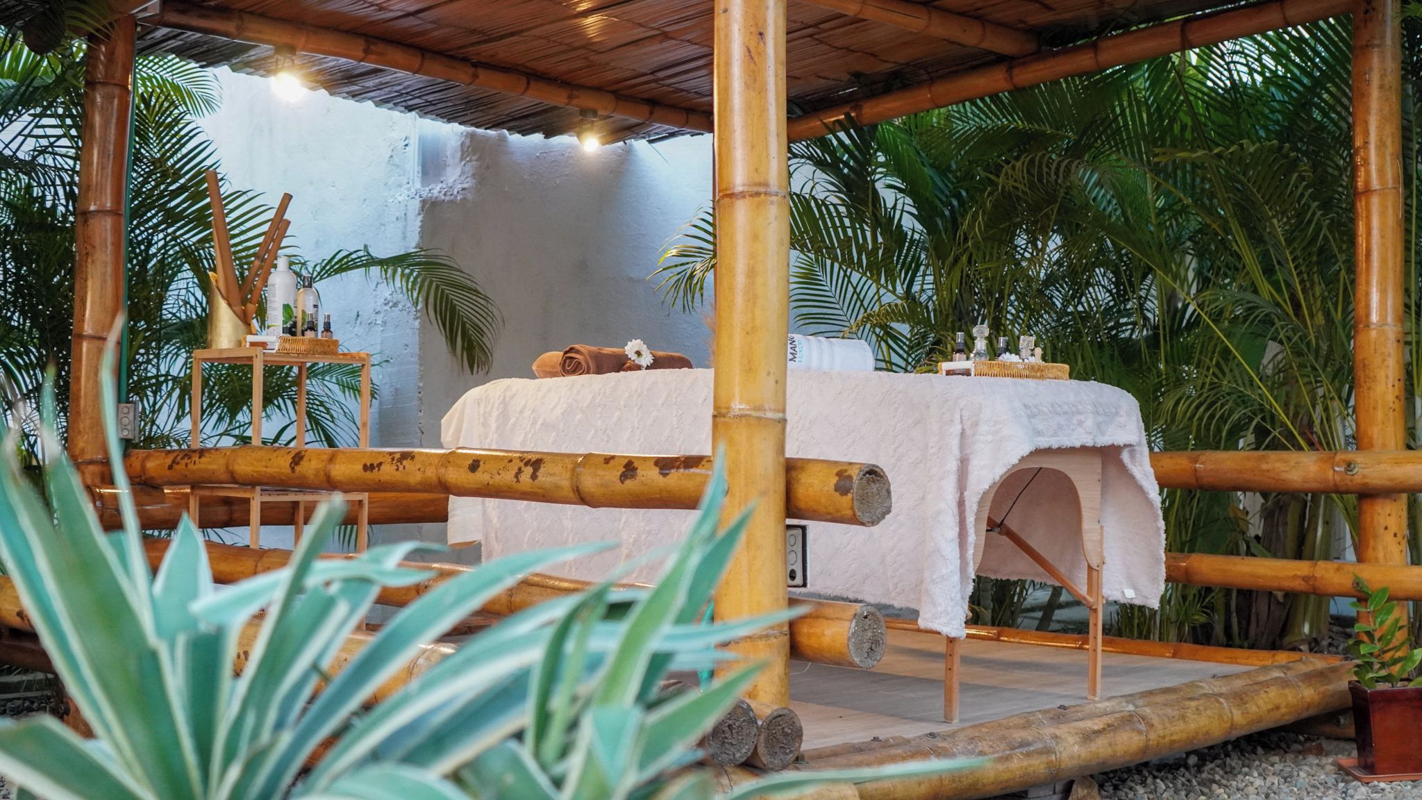 A bamboo-framed outdoor seating area with a table covered in a white cloth, surrounded by tropical plants.