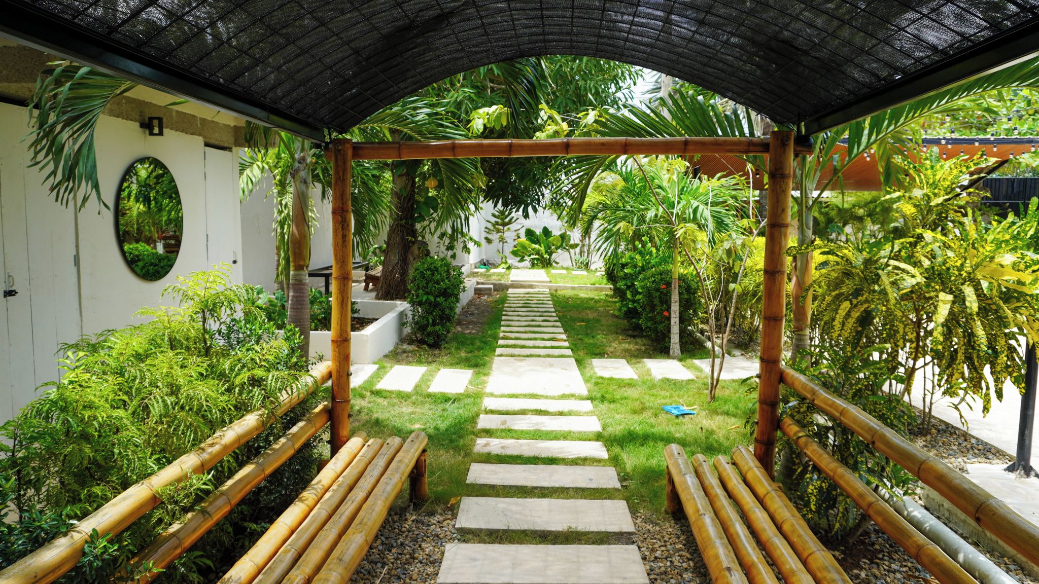 A covered garden pathway with benches, surrounded by lush greenery and a clear sky.