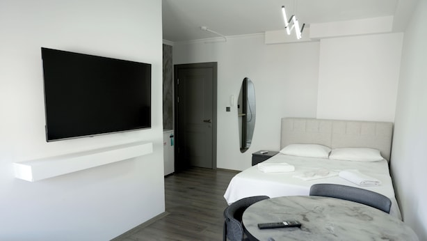 Quarto de hotel moderno com televisão de tela plana na parede, cama com cabeceira cinza, mesa redonda pequena com um controle remoto e uma cadeira.