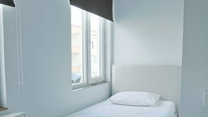 Quarto individual com uma cama, janela, radiador e cabeceira branca.
